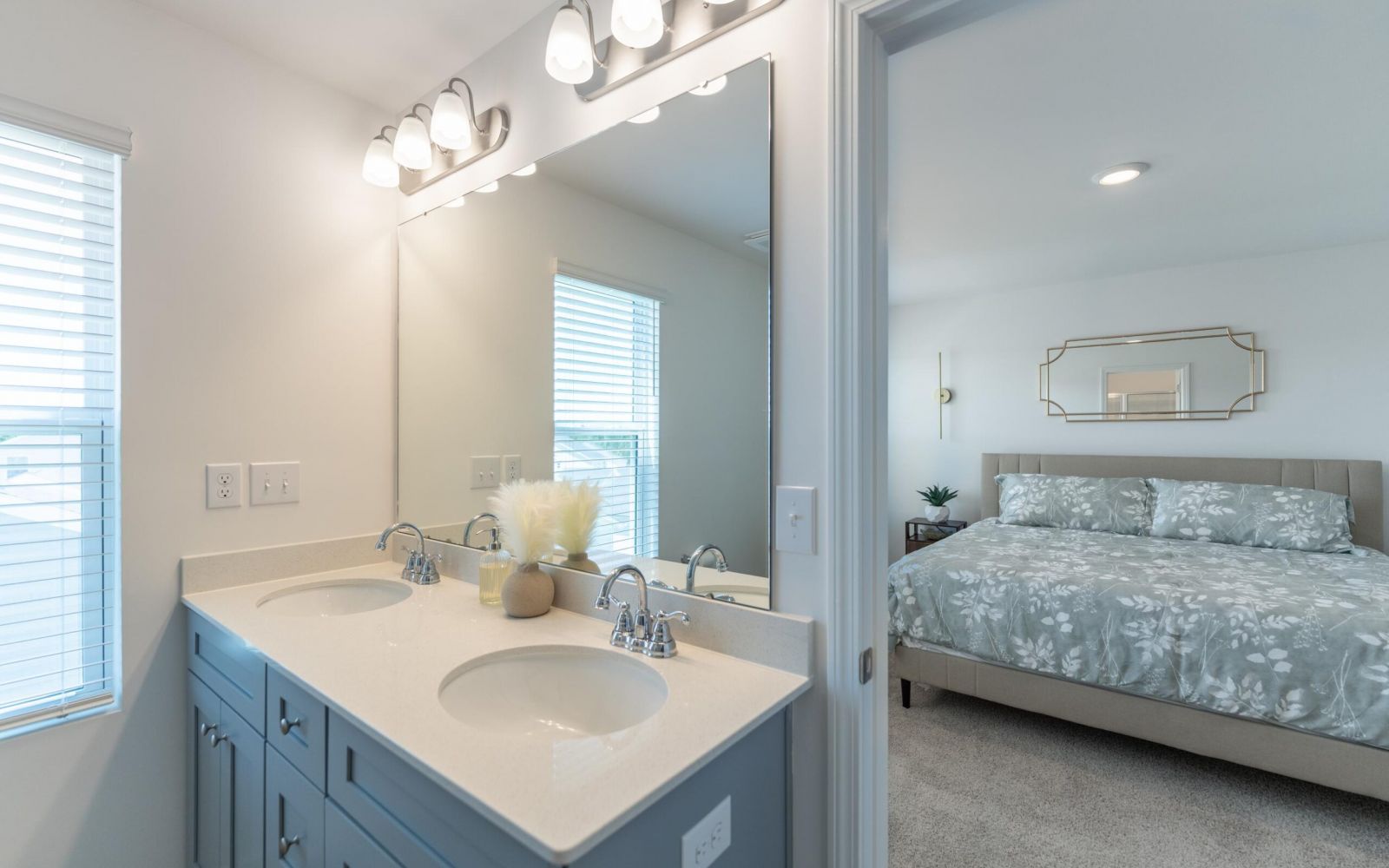 The Bend at Cramerton Mills modern bathroom with double sinks and large mirror, connected to a bright bedroom with a made bed.
