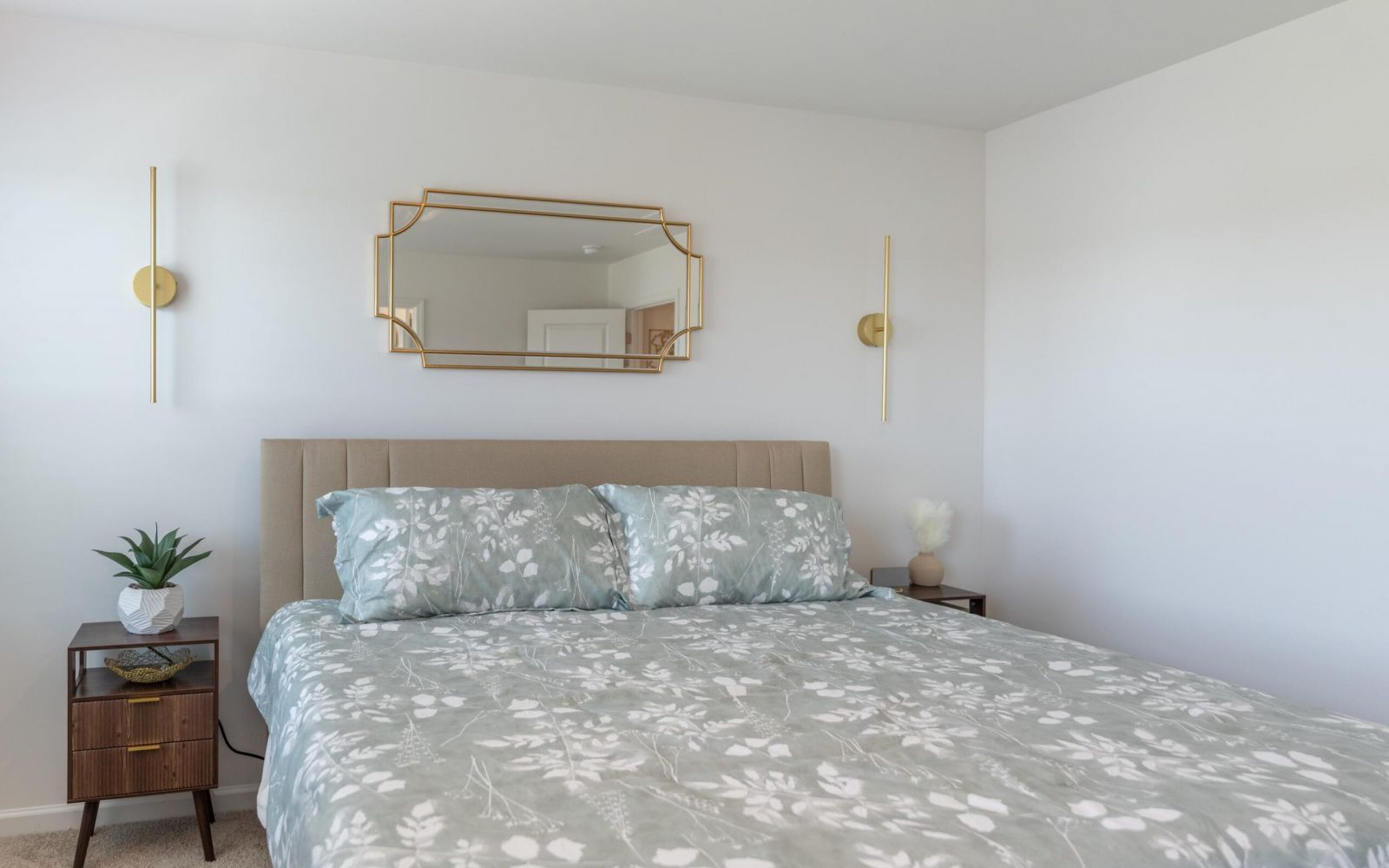 The Bend at Cramerton Mills a neatly made bed with floral bedding, a gold-framed mirror, and a wall lamp in a bright bedroom.